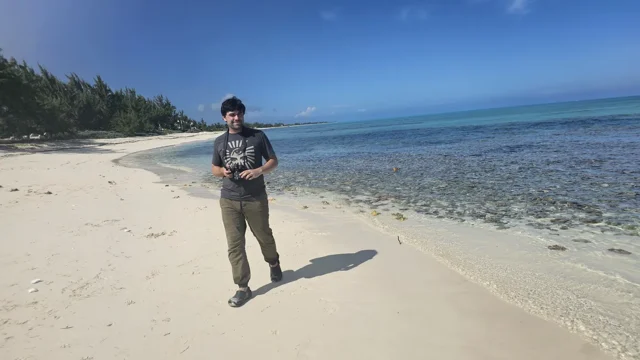 Ryan holding a drone controller on a sandy beach beside blue water.