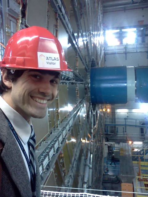 Ryan wearing a red ATLAS visitor hard hat at CERN, with the detector hall behind him.