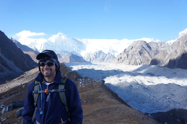 A hiker in a blue jacket and sunglasses, with a glacier and snowy mountain peaks behind him.