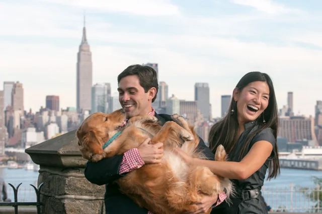 A couple holds a golden dog on a rooftop, with the Manhattan skyline behind them.